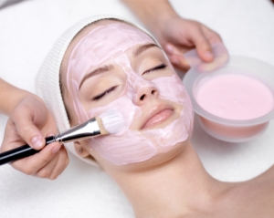 Esthetician applying a light pink colored serum to woman's face using a brush.