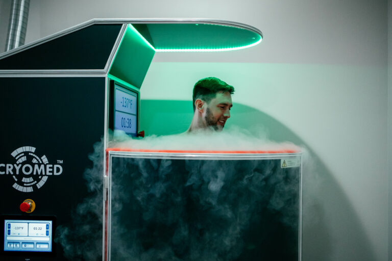 Man standing inside Cryotherapy chamber with a green light overhead and smoke overflowing from the chamber.